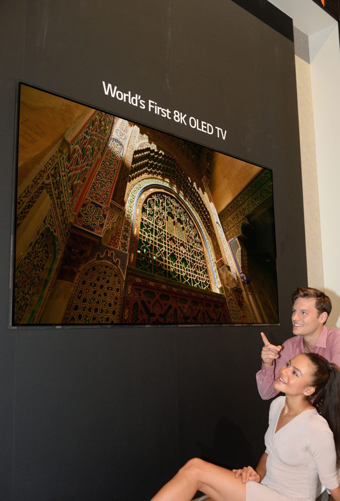 Another view of the World’s First 8K OLED TV display at IFA 2018, with a male and female model sitting below and pointing up to the screen