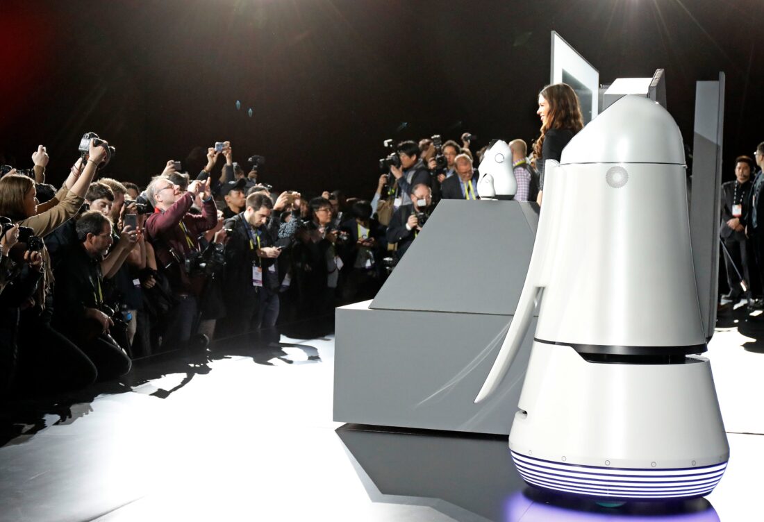 Side view of a female model posing with LG CLOi Robots on stage while reporters take pictures at LG's CES 2017 Press Conference.