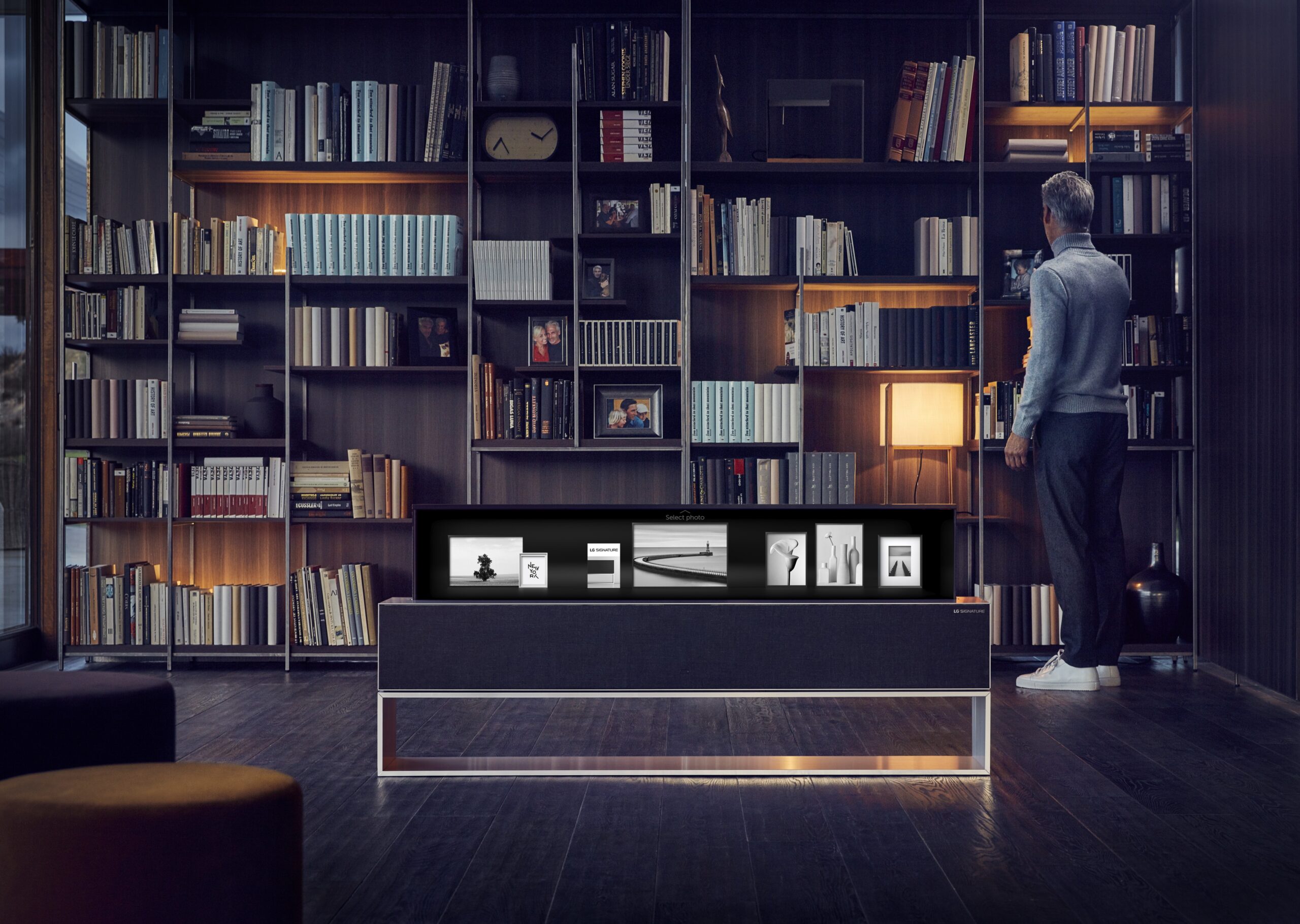 A man is browsing his bookshelf, an LG SIGNATURE OLED TV R enhances the environment by displaying photos on its screen