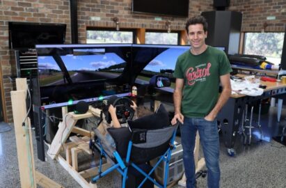 Rick Kelly poses and in front of his new racing simulator as his son uses it behind him