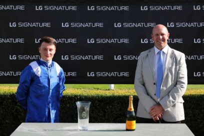 A winning jockey from the Qatar Goodwood Festival is presented with a glass trophy and a bottle of champagne in front of an LG SIGNATURE-branded sign
