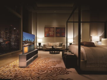 Left-side view of LG SIGNATURE OLED R inside a modern bedroom in Full View mode, its on-screen image blending into the night skyline seen from the large window behind