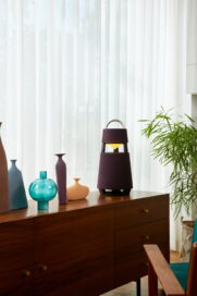 LG XBOOM 360 speaker blending with decorative objects on a wooden cabinet, its light matching the warm sunlight from outside