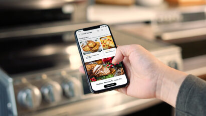 A hand holding a smartphone displaying recipes on the LG ThinQ Recipe app.