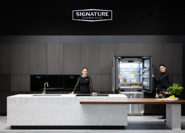A woman is standing behind the kitchen island and a man is holding the Signature Kitchen Suite built-in 36-inch French Door refrigerator door at LG booth