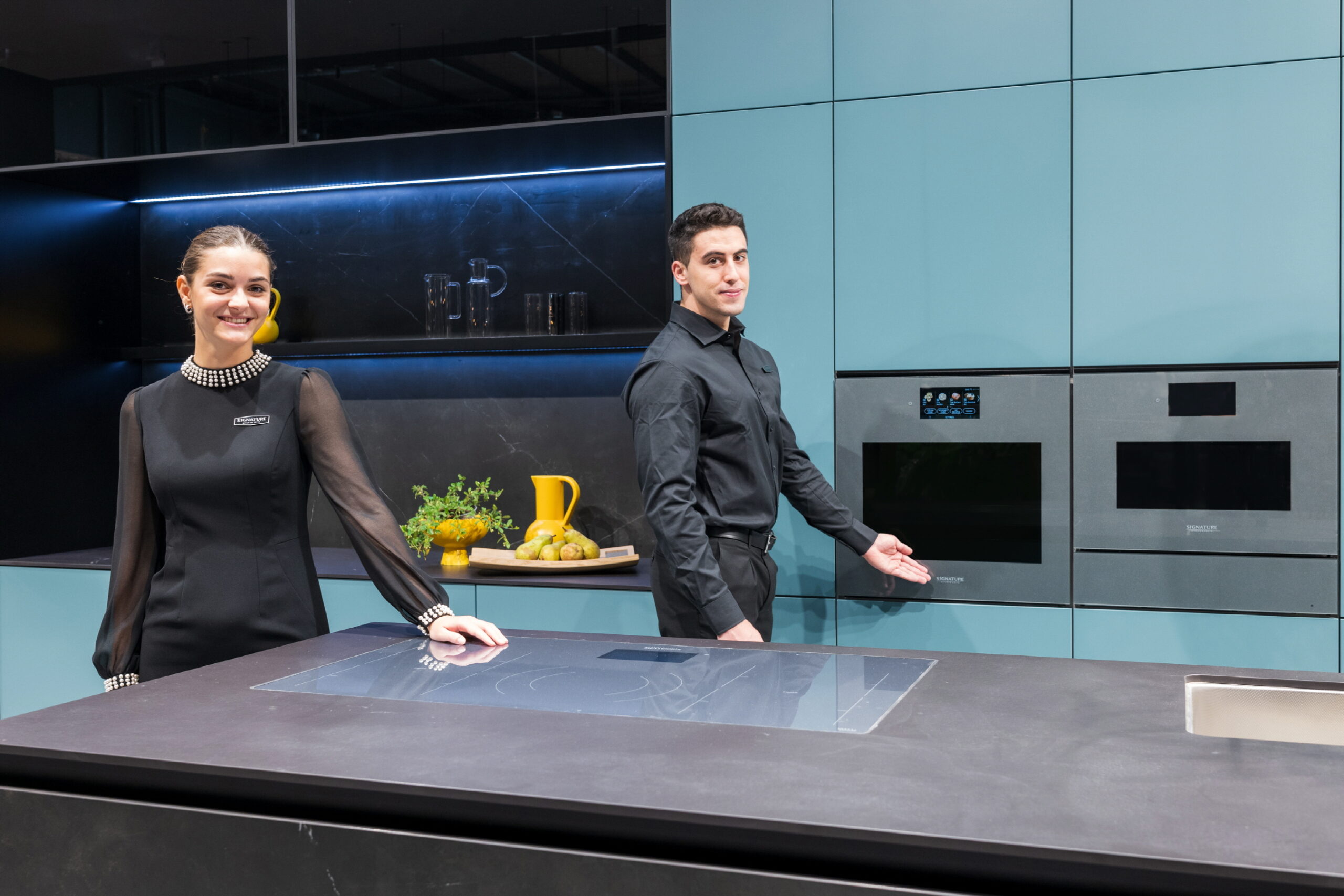 A woman on the left is standing next to the Signature Kitchen Suite 36-inch induction and a man is indicating the Signature Kitchen Suite 24-inch steam oven