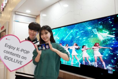 A man and woman posing in front of an LG Smart TV displaying a K-pop group’s performance to celebrate more K-culture content coming to LG’s webOS platform.