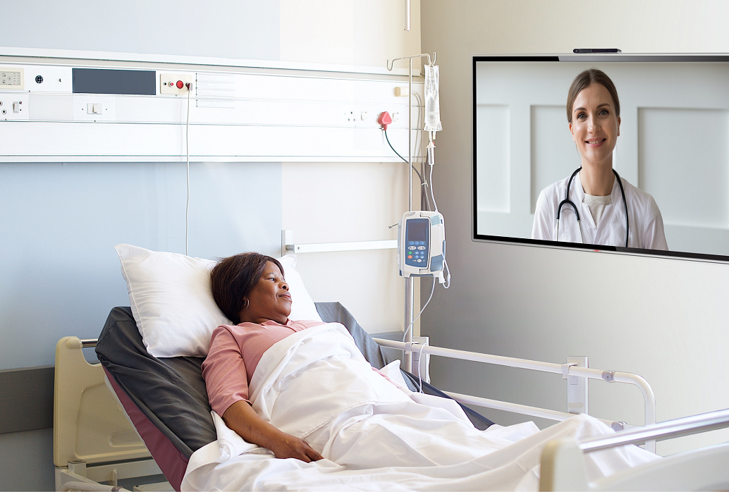 A patient who is resting in a bed is having a video chat with her doctor with the help of LG healthcare TV installed on the wall.