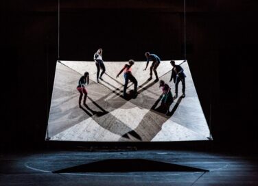 Dancers of the National Choreographic Center of Grenoble balancing on a rectangular platform hanging in the air during the He Who Falls performance