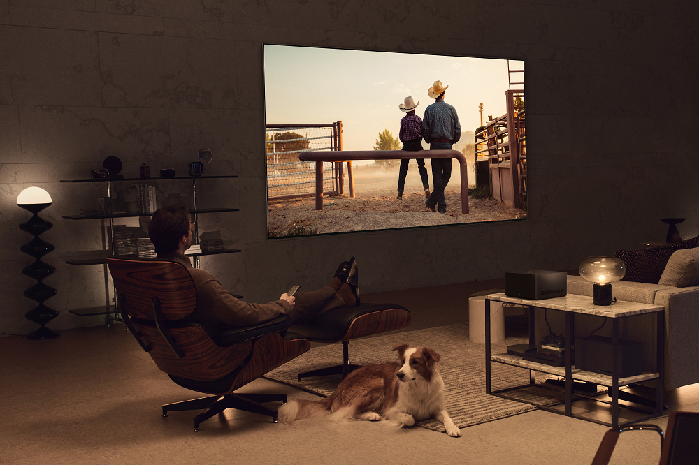 A man watching a TV show on the new, wall-mounted LG OLED M3 as he relaxes on his living room chair beside a dog