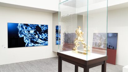 A gold sculpture inside a glass display case with an LG OLED TV and Objet Easel displaying artwork in the background