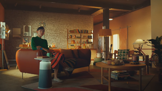 A woman sitting on the living room sofa while using LG PuriCare Aero Furniture in Crème Grey as a table for her mug and book. 