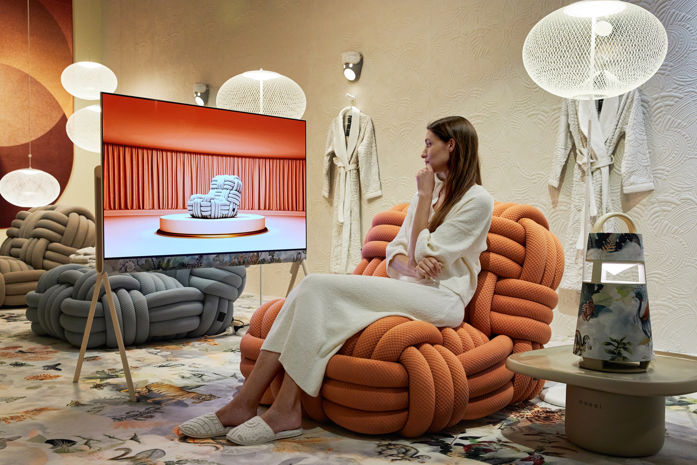 A woman sitting on a stylish piece of Moooi furniture as she watches content on LG OLED Objet Collection Posé and sits next to a limited-edition LG XBOOM 360 portable speaker