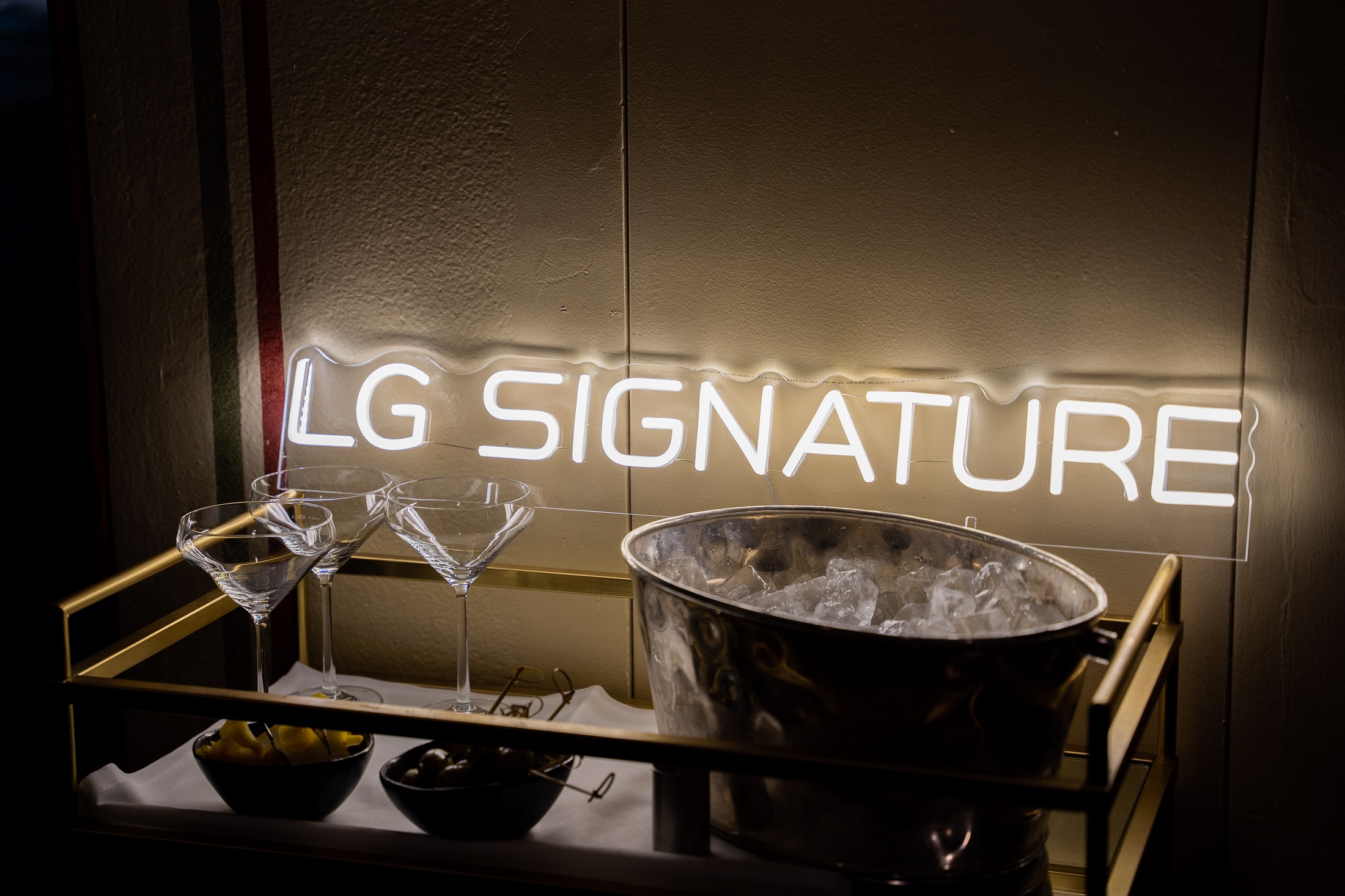 A photo of a LG SIGNATURE sign placed behind the champagne glasses, a cup of olives and a bucket of ice