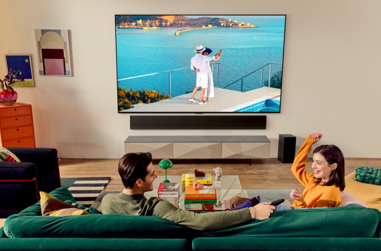 A man and a woman sitting on a green sofa and having fun while watching a TV displaying a couple dancing