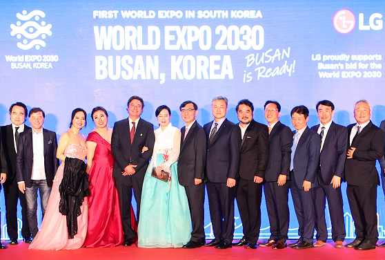 A photo of opera-related actors and staff posing on the podium with a digital screen at the back displaying the phrase "WORLD EXPO 2030 BUSAN, KOREA"