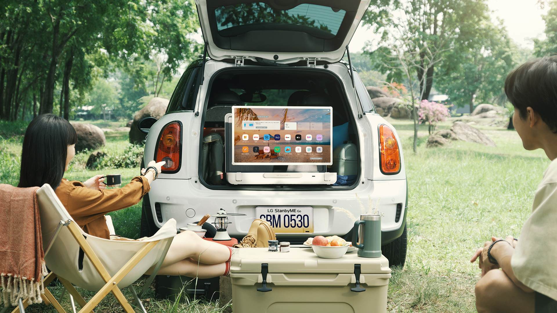 A young couple on a camping trip using LG StanbyME Go which is placed in the open trunk of their car