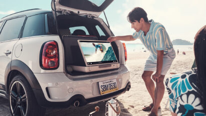 A couple watching a surfing video on the beach with LG StanbyME Go positioned in the open trunk of their car