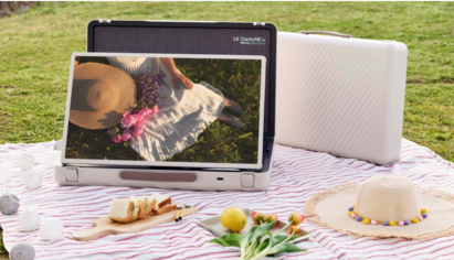 A blanket is spread out on a grassy lawn for a picnic with the LG StanbyME Go model in landscape mode displaying the image of a woman