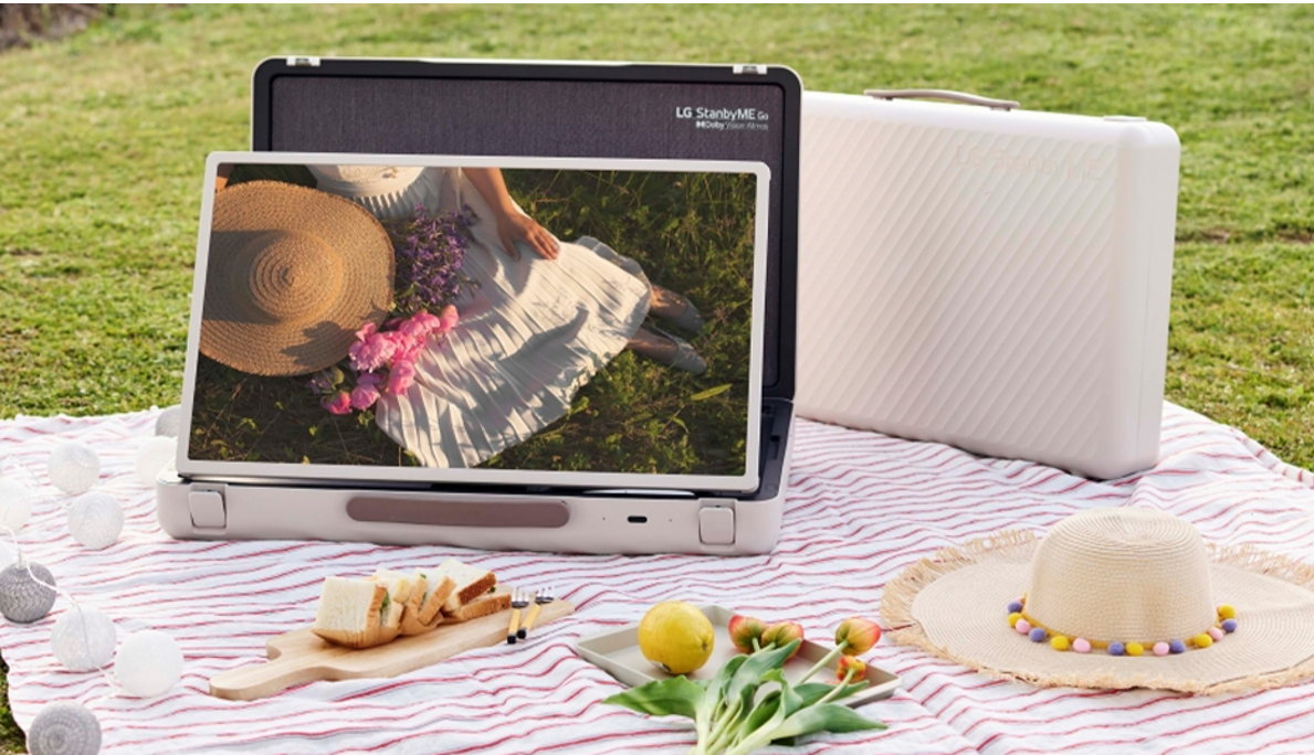 A blanket is spread out on a grassy lawn for a picnic with the LG StanbyME Go model in landscape mode displaying the image of a woman