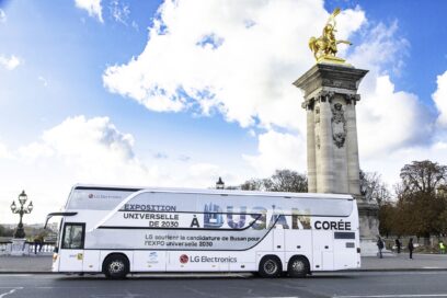 A photo of a big LG bus with the words 'A Busan Coree' passing by a statue in the city