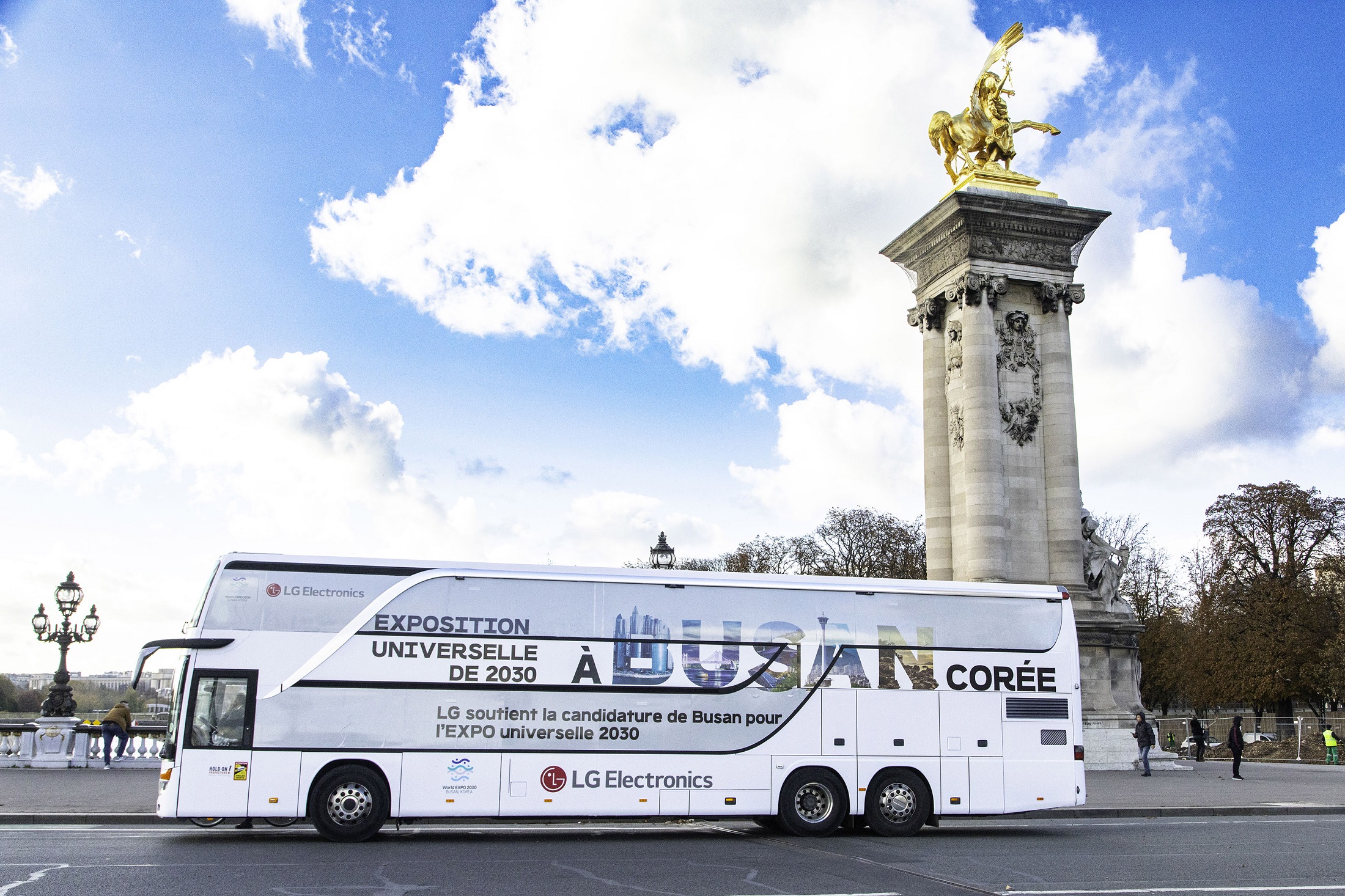 A photo of a big LG bus with the words 'A Busan Coree' passing by a statue in the city