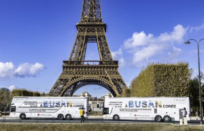 A photo of two LG Buses with the words 'A Busan Coree' closely passing by the Eiffel Tower