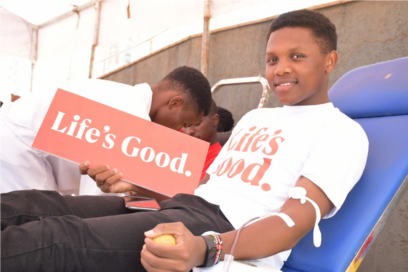 A blood donor wearing Life's Good T-shirt posing for a photo while holding a panel that also reads 