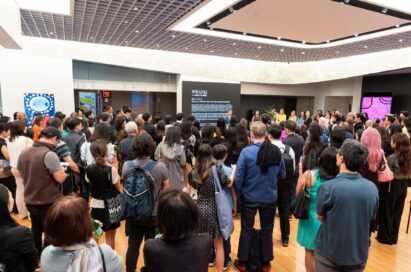 A crowd of people are gathered to listen to a presentation by a man standing in front of a wall of information titled “Whanki.”