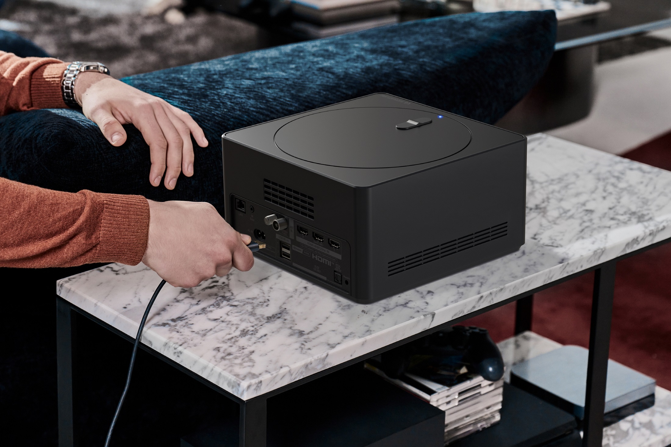 A man is connecting a cable to a Zero Connect Box, which sits on a side table that hides video games and controllers underneath