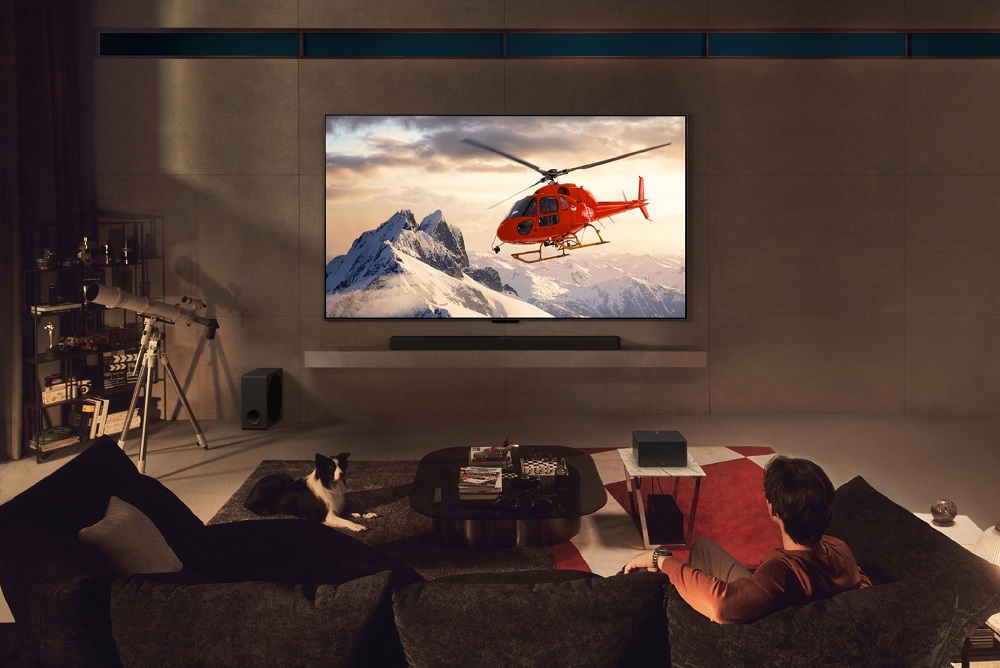 A photo of a man watching a helicopter on the screen of a wall-mounted LG OLED M4, with a wireless Zero Connect Box sitting on a small table in the living space