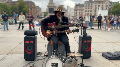 A street artist in the UK sits in a city square between two LG XBOOM speakers while playing a multi-neck guitar and singing into a microphone