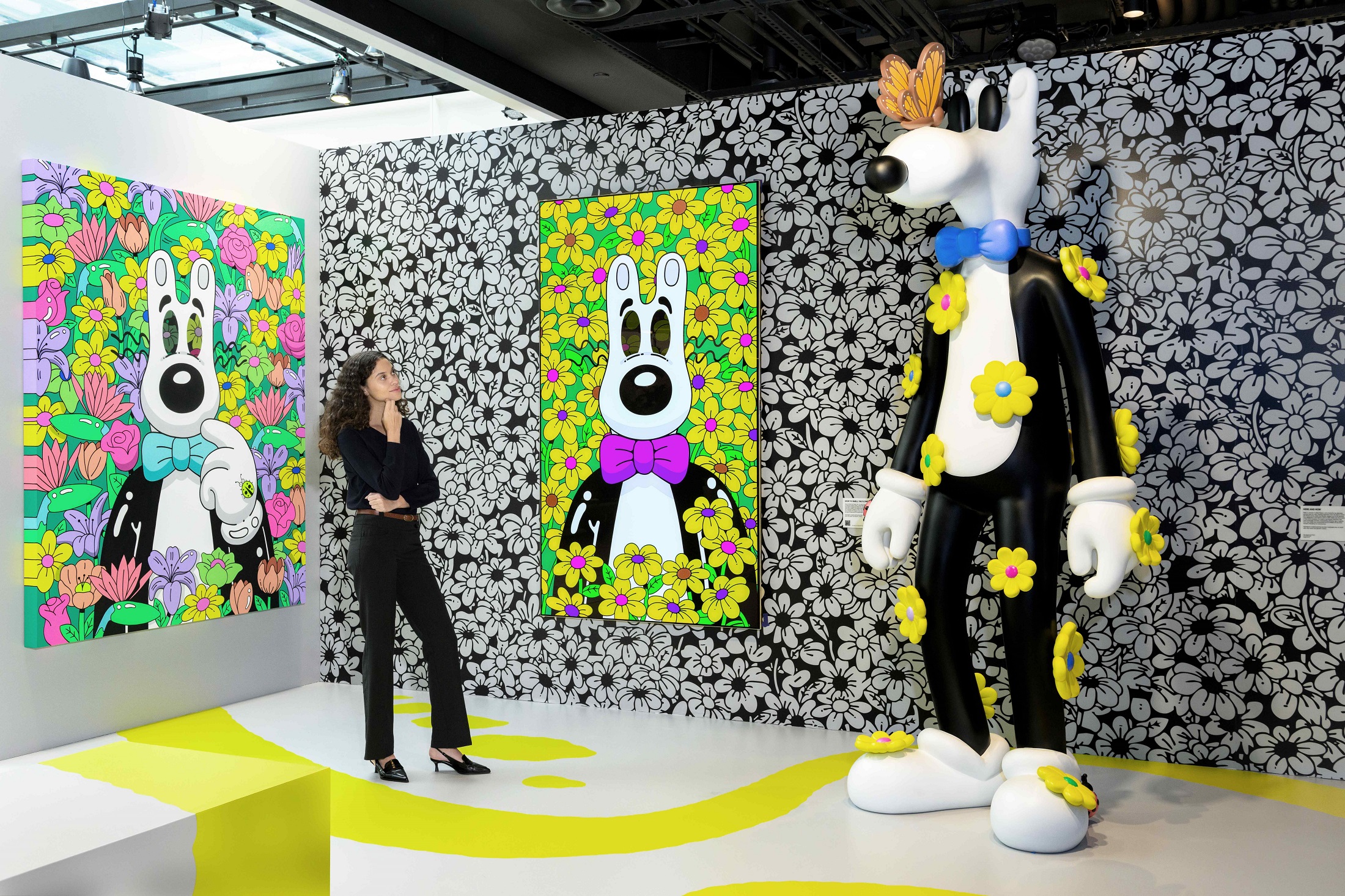 A woman is exploring Steven Harrington's vibrant floral-themed artwork and looking at a large playful sculpture of Mello in an immersive exhibition space.